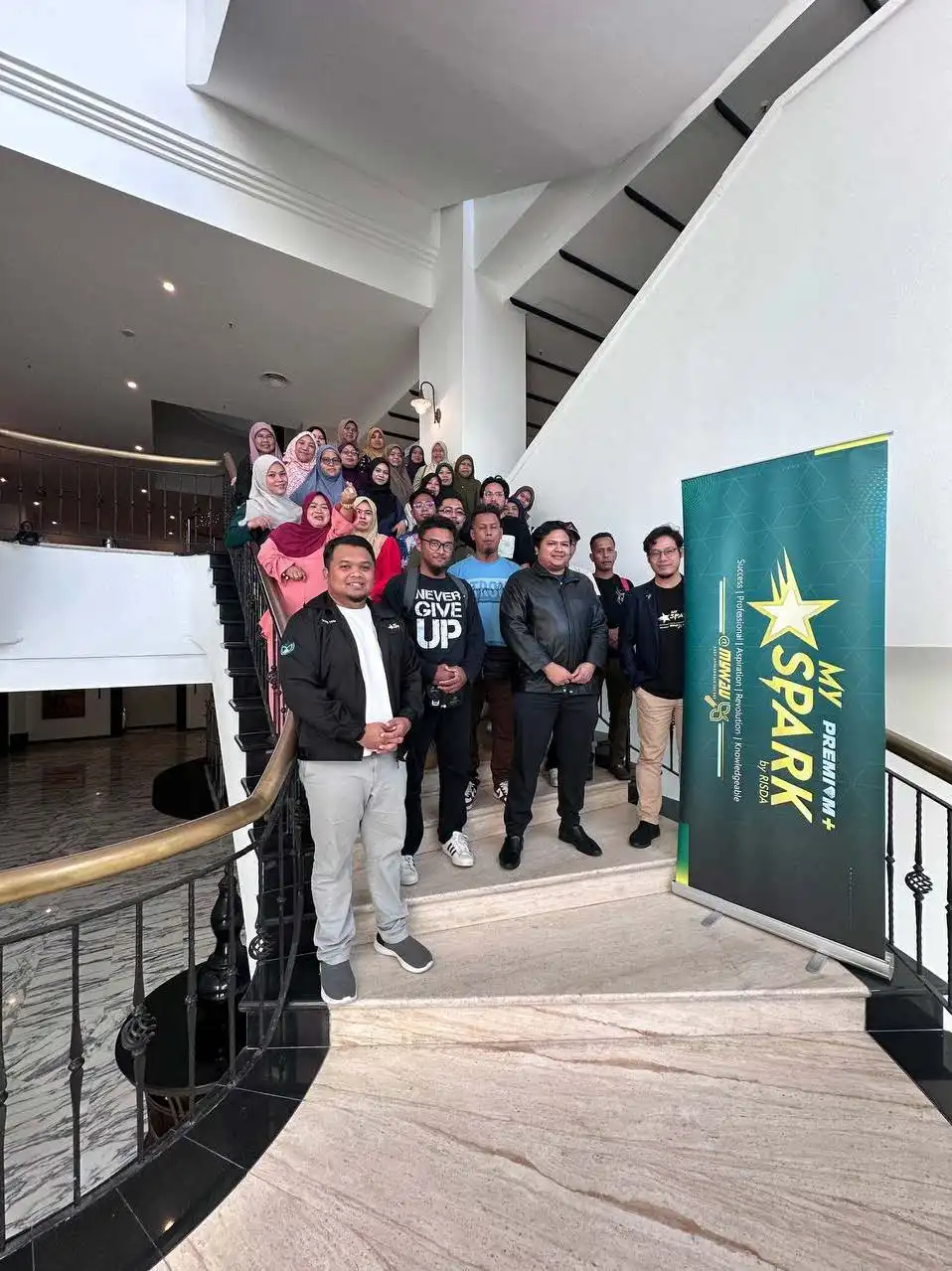 A group of people, gathered for a coaching session, stand on a staircase next to a My Spark banner in a building with white walls and a black railing. CK Media
