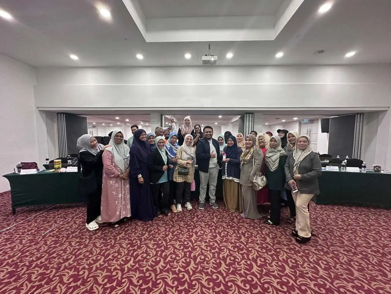 A group of people, some wearing hijabs, pose for a photo in a carpeted conference room where coaching sessions are held, with a long table in the background. CK Media
