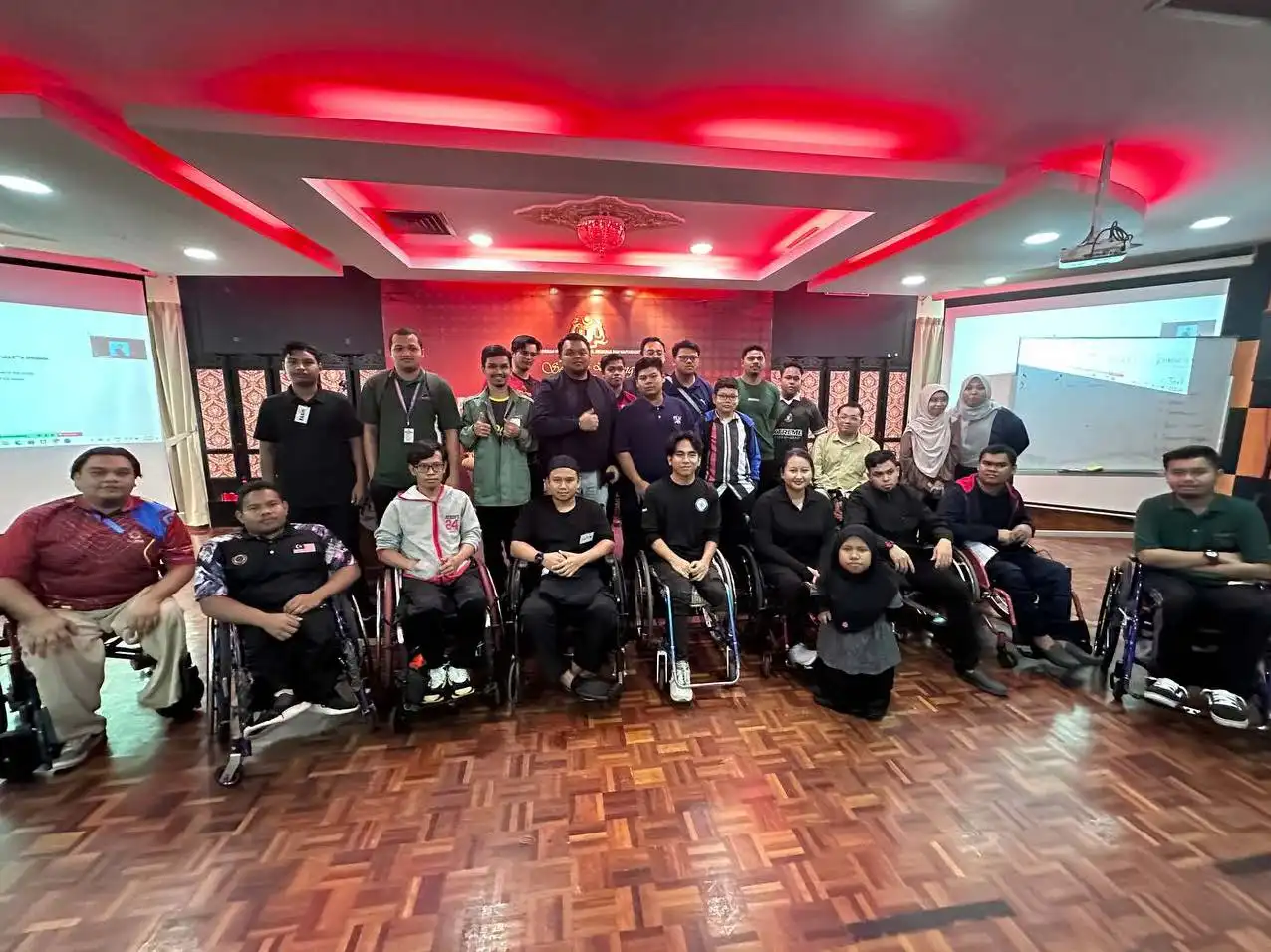 A group of people, some using wheelchairs, pose for a photo in a room with a red-lit ceiling and wooden floor. Presentation screens in the background suggest an empowering coaching session. CK Media