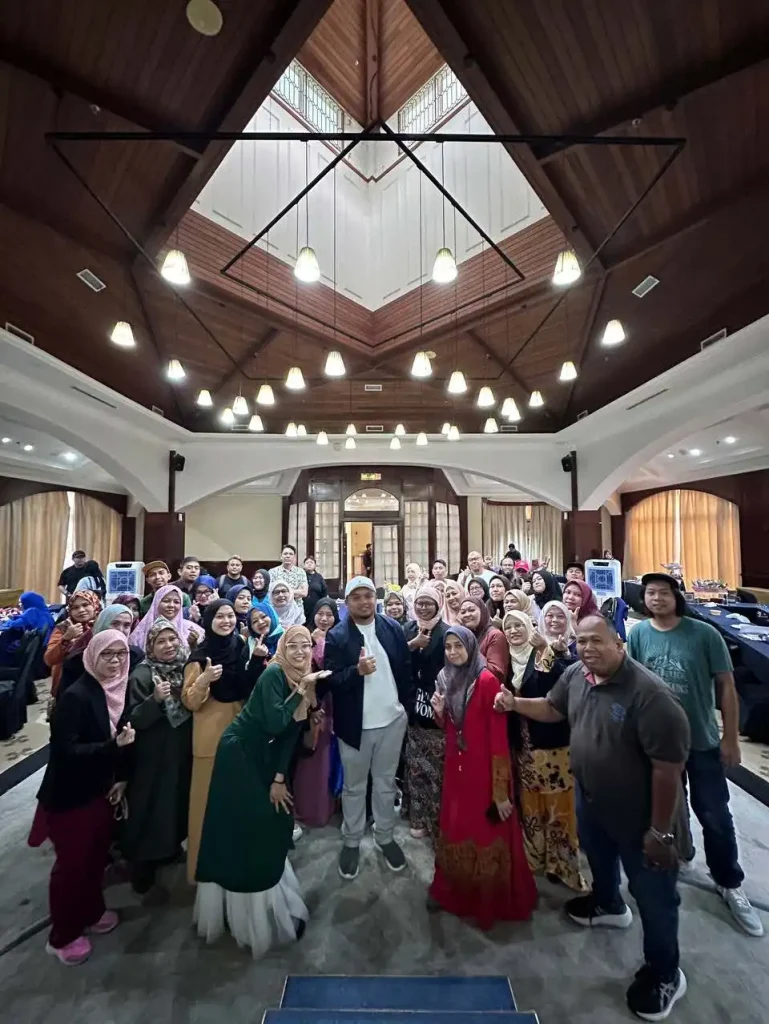 A group of people, united by recent training sessions, stand together in a large room with a high ceiling and hanging lights, proudly posing for a group photo. CK Media