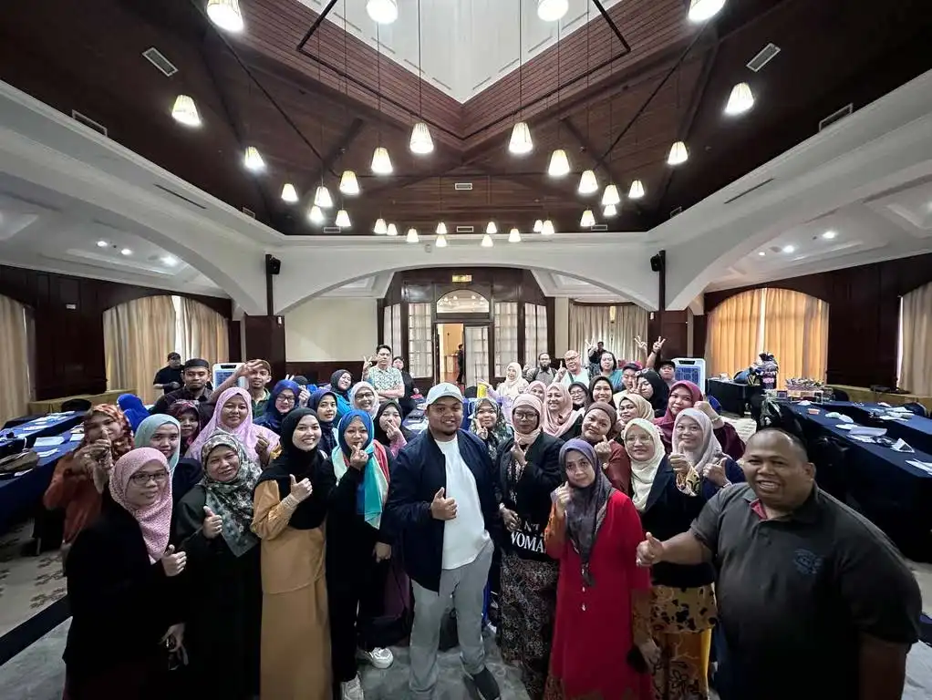 A large group of people, some wearing headscarves and others in casual clothing, stand together in a spacious room with high ceilings, smiling and giving thumbs up after an inspiring coaching session. CK Media